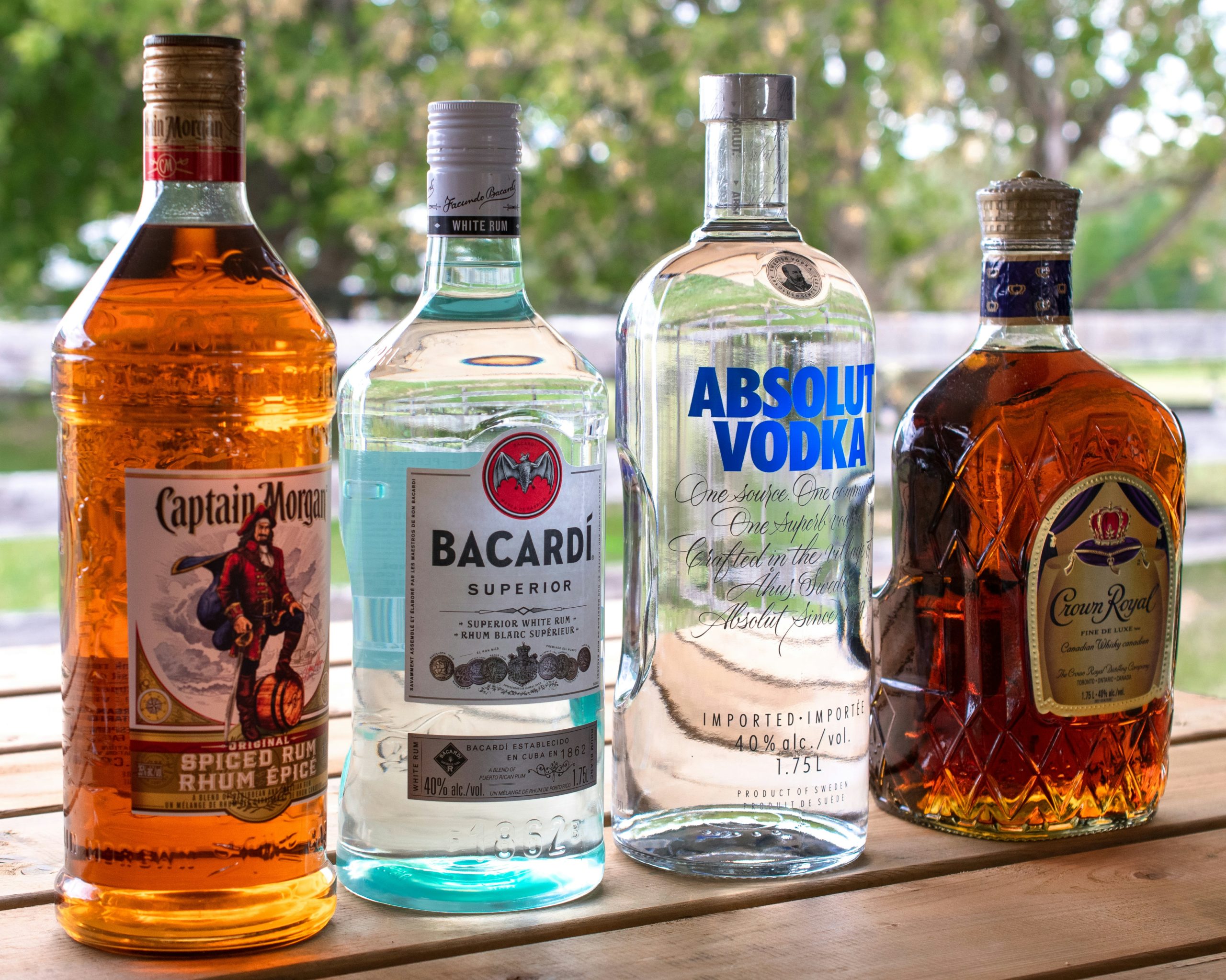 Четыре бутылки алкоголя: Captain Morgan, Bacardi, Absolut Vodka и Crown Royal, стоящие на деревянной поверхности.